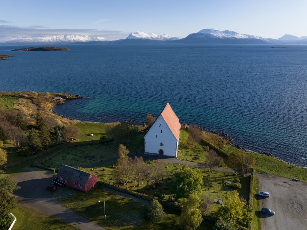 Kyrkjemidlar til Trondenes: En hvit kirke med rødt tak står nær strandlinjen, omgitt av grøntområder, med fjell og vann i bakgrunnen under en klar himmel.