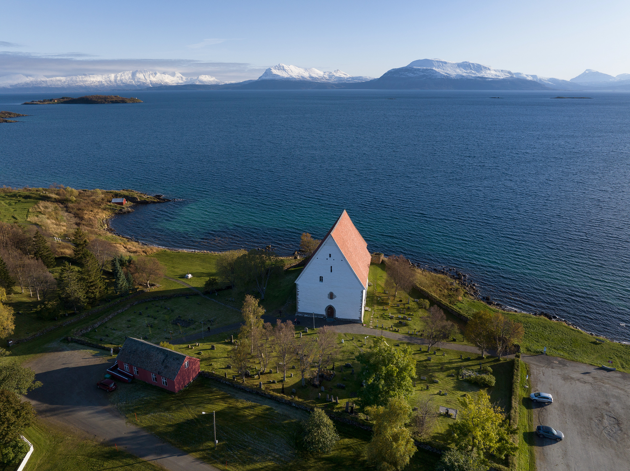 Kyrkjemidlar til Trondenes: En hvit kirke med rødt tak står nær strandlinjen, omgitt av grøntområder, med fjell og vann i bakgrunnen under en klar himmel.