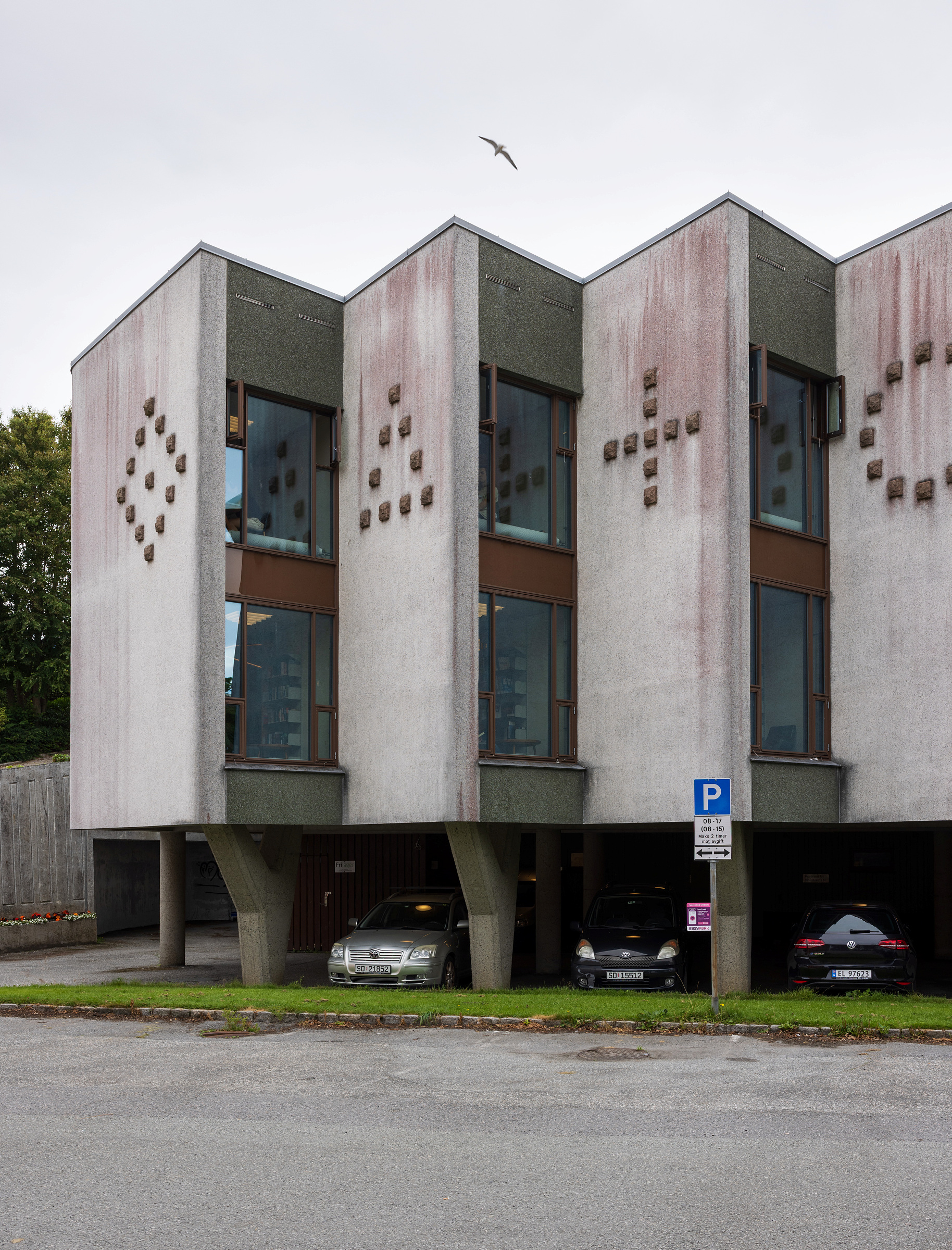 Tre leilighetsenheter i betong med geometrisk veggdesign står over parkeringsplasser, med en bil parkert under hver enhet og en fugl som flyr over hodet.