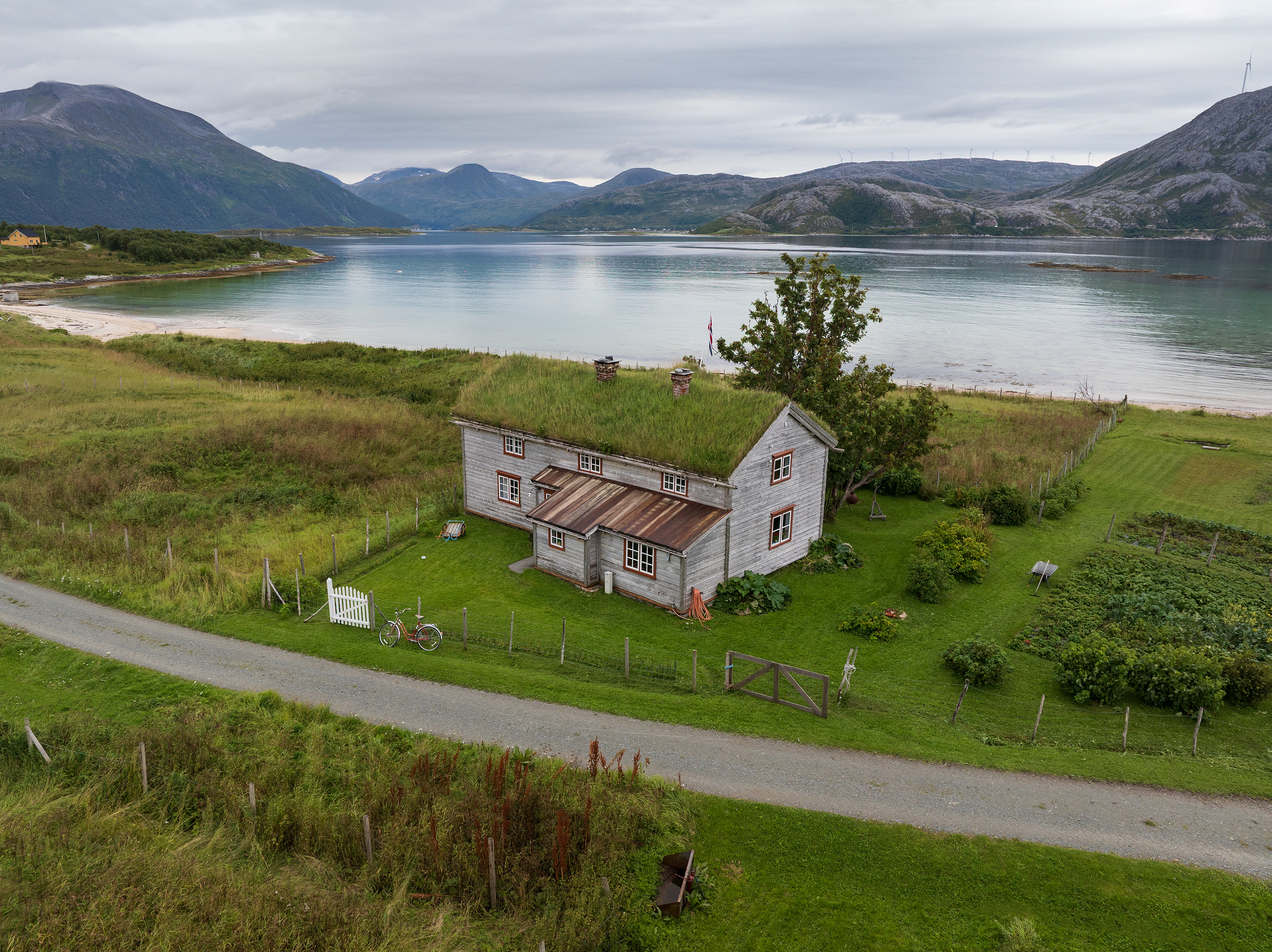 Et tradisjonelt trehus med gresskledd tak ligger ved en grusvei, nær en stille innsjø og omgitt av fjell under en overskyet himmel.