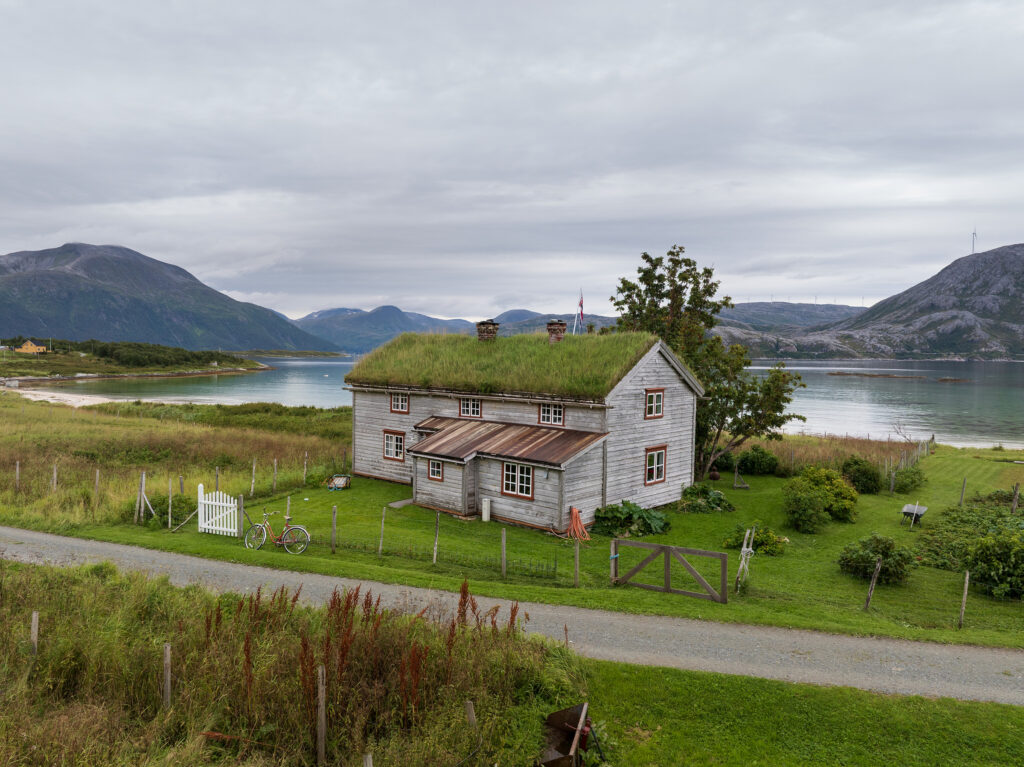 Et trehus med gresskledd tak ligger ved en innsjø og fjell, omgitt av en hage og et gjerde på en overskyet dag.