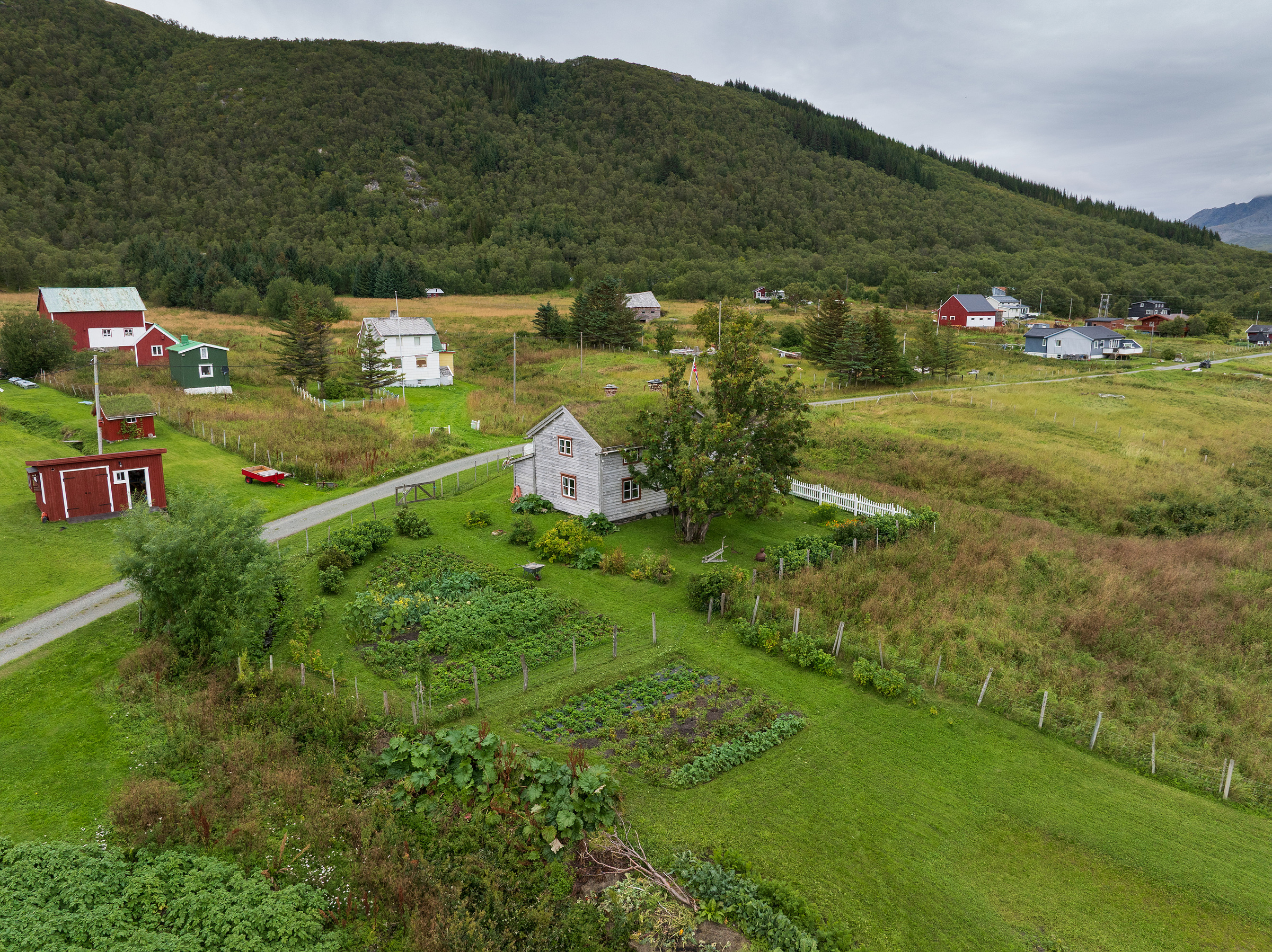 Et lite steinhus med grønnsakshage er omgitt av gressletter, trær og spredte hus ved foten av en skogkledd ås under en overskyet himmel.