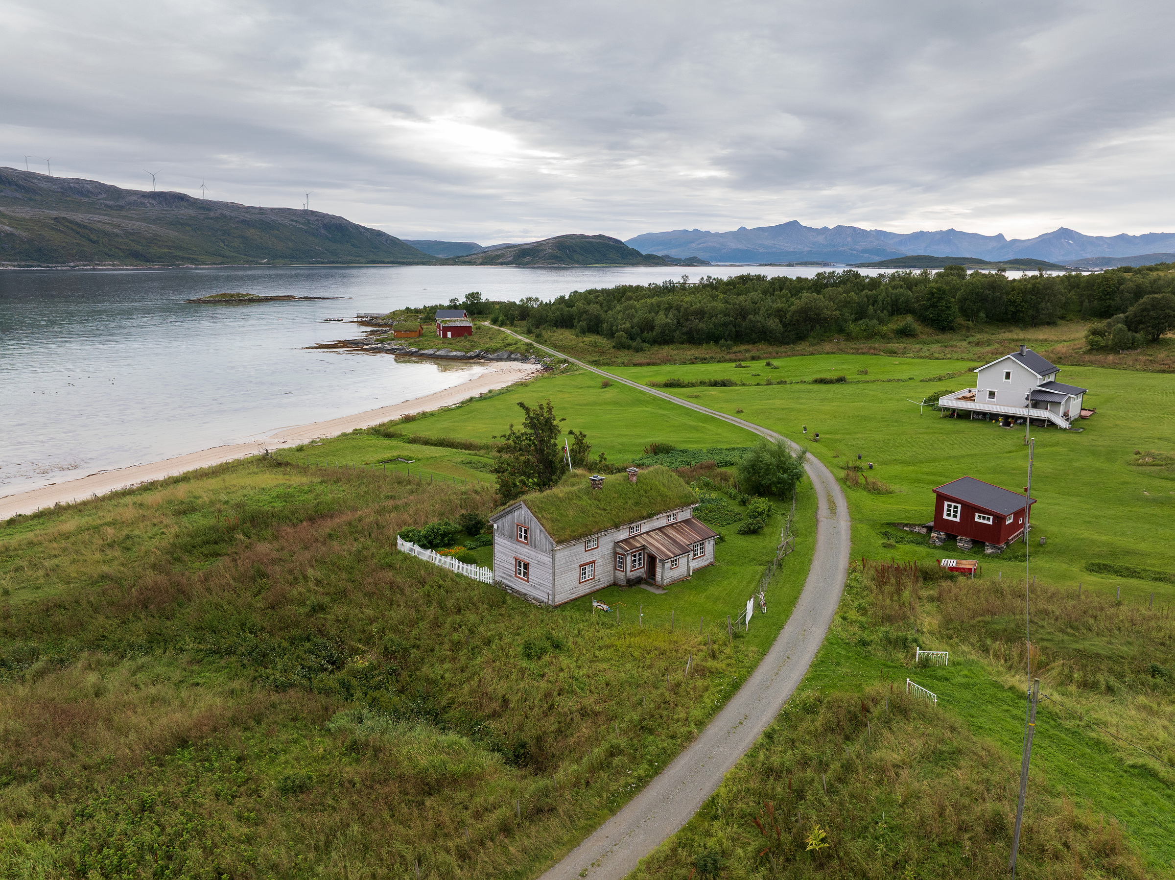 Et landlig landskap med en svingete vei, spredte hus, grønne åkrer, en strandlinje og fjell i bakgrunnen under en overskyet himmel.