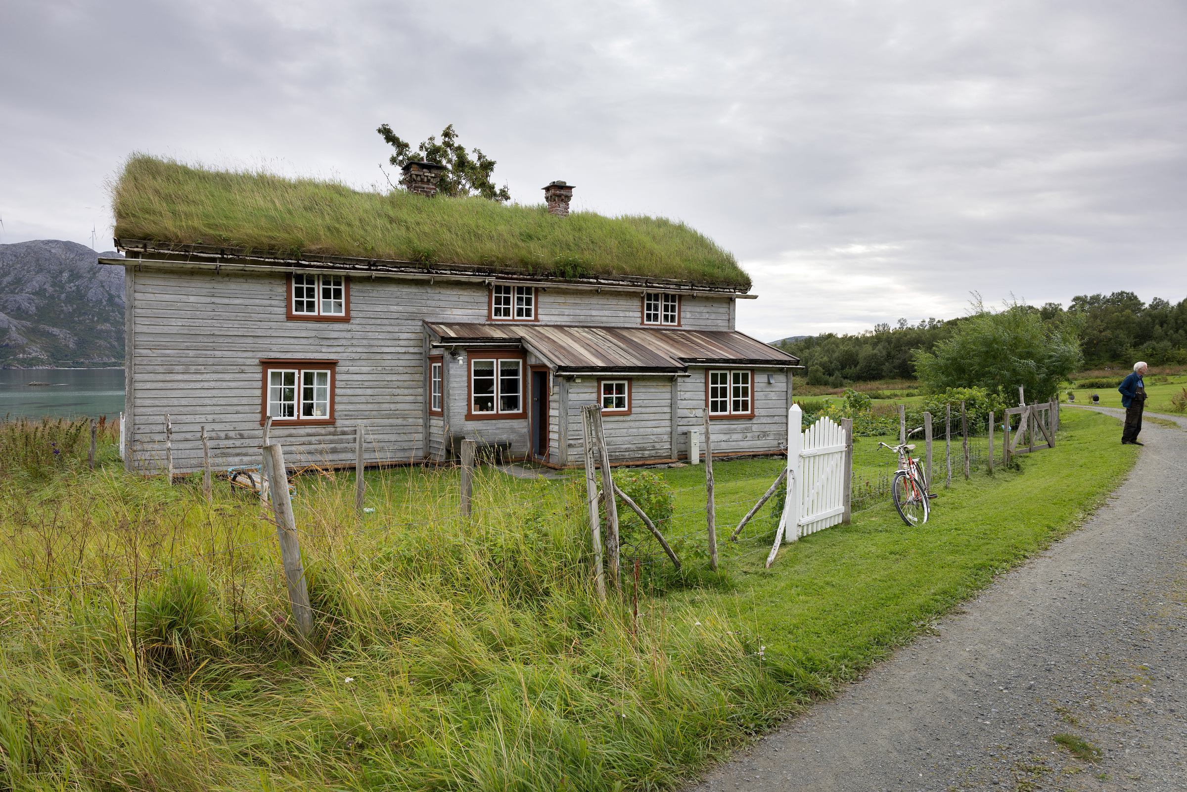 Et forvitret trehus med gresstak står bak en hvit port og et gjerde nær en grussti; en sykkel står lent mot gjerdet, og en person står like ved.