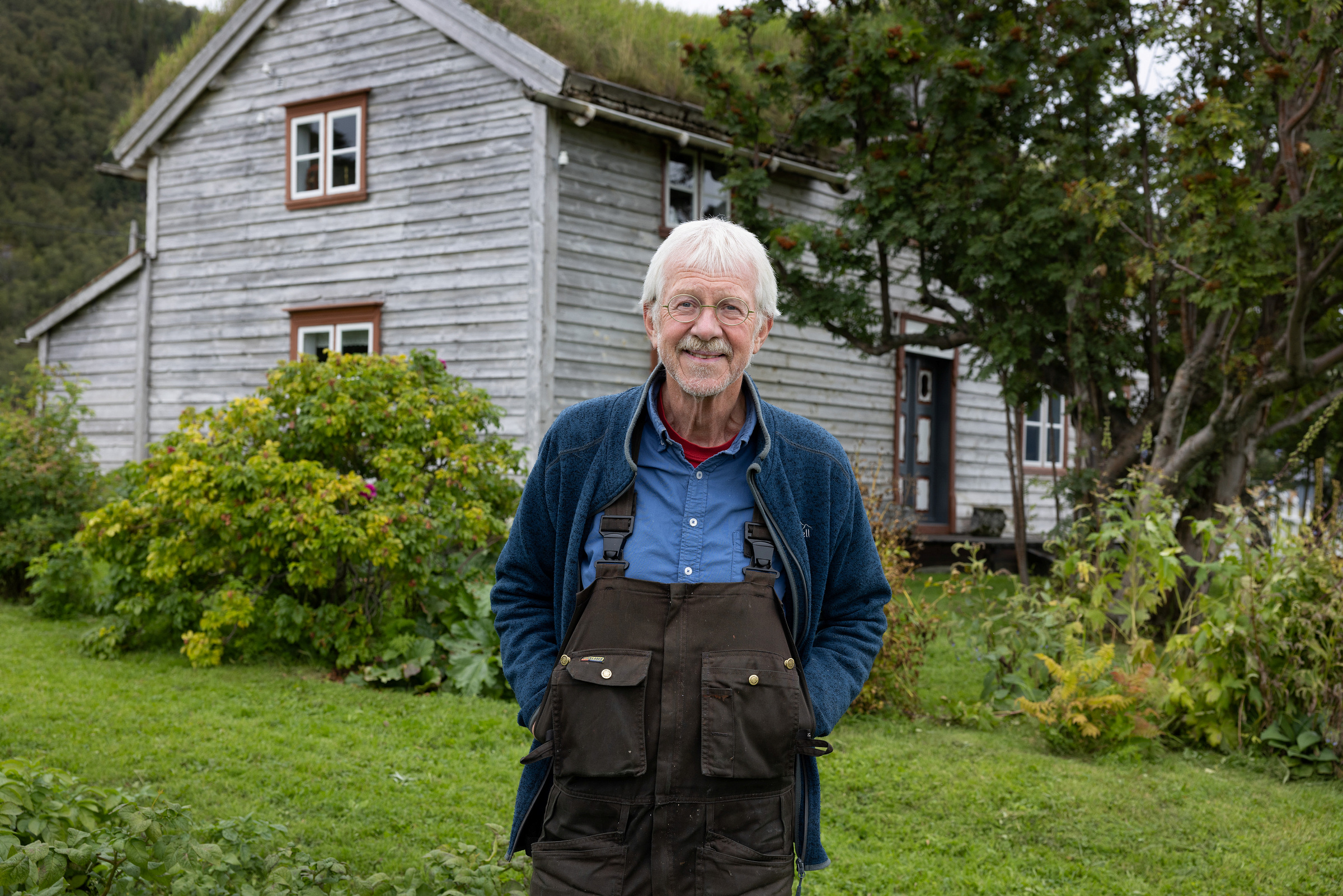 En eldre mann med hvitt hår og briller står foran et forvitret trehus, iført blå skjorte, jakke og brun kjeledress, omgitt av grønne planter.