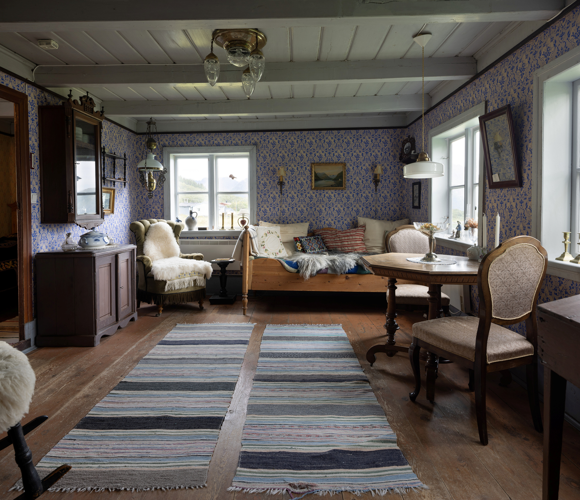 En koselig stue i vintage-stil med mønstret tapet, tremøbler, stripete tepper og naturlig lys fra flere vinduer.
