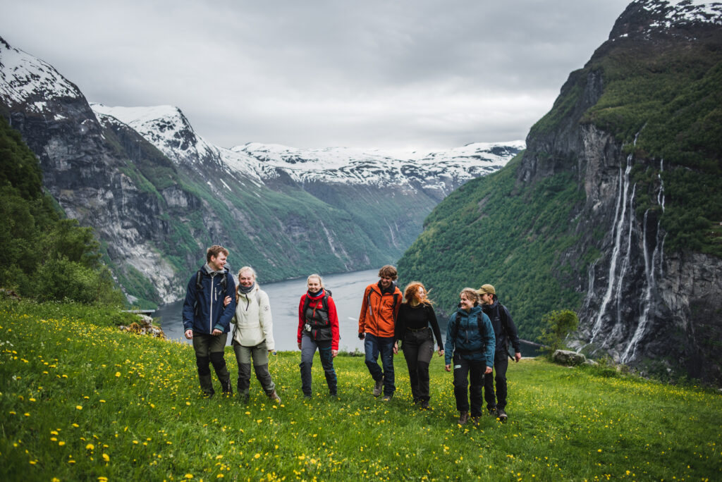7 ungdommer står i en grønn dal, og ser mot kamera. Det er en storslått fjord i bakgrunn og flere fossefall.