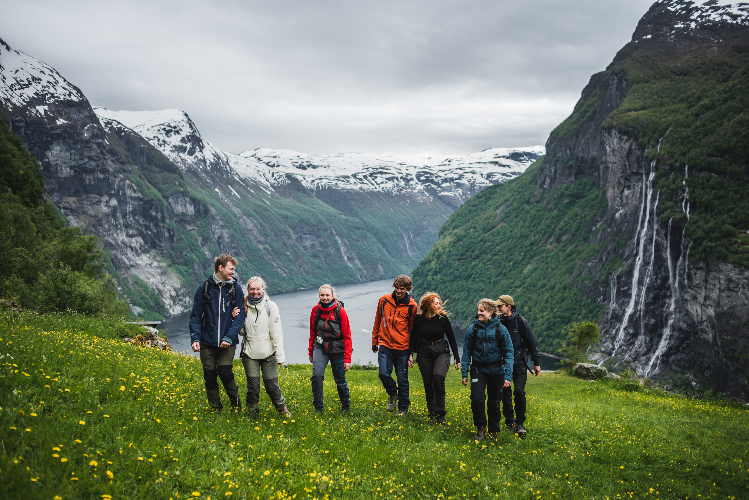 7 ungdommer står i en grønn dal, og ser mot kamera. Det er en storslått fjord i bakgrunn og flere fossefall.