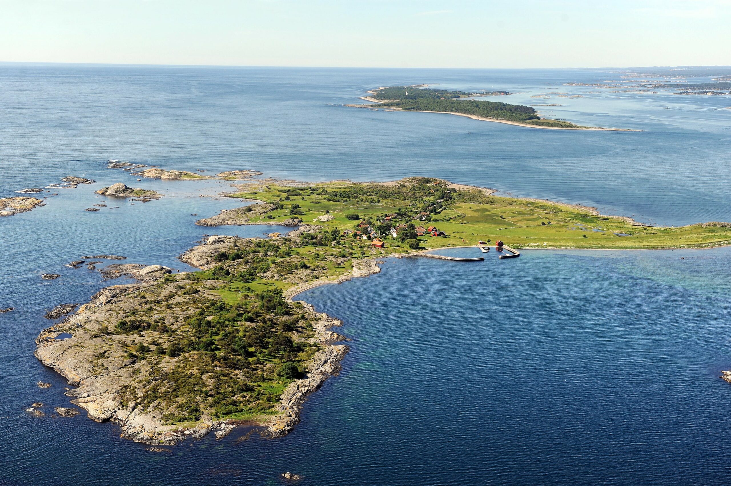 Flyfoto av en liten, steinete øy med spredte hus, grønne åkrer og en brygge, omgitt av stille, blått vann.
