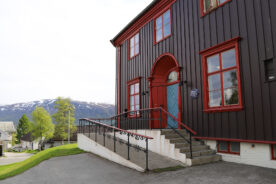 En stor, mørkebrun trebygning med røde kanter, trapper og en rullestolrampe foran, med trær og snødekte fjell som bakgrunn.