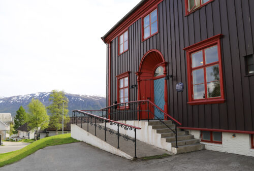 En stor, mørkebrun trebygning med røde kanter, trapper og en rullestolrampe foran, med trær og snødekte fjell som bakgrunn.