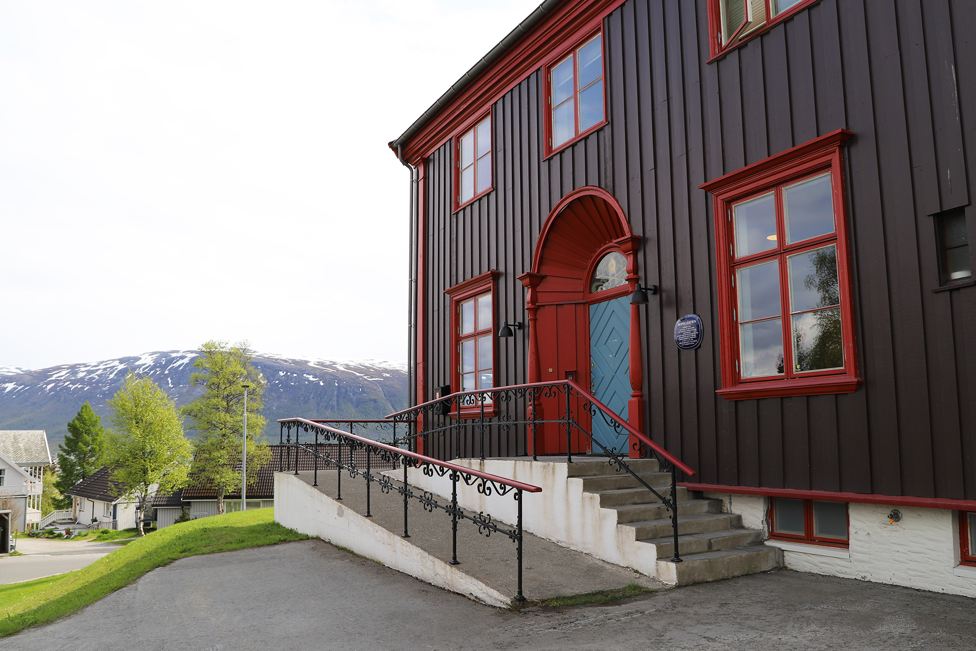 En stor, mørkebrun trebygning med røde kanter, trapper og en rullestolrampe foran, med trær og snødekte fjell som bakgrunn.