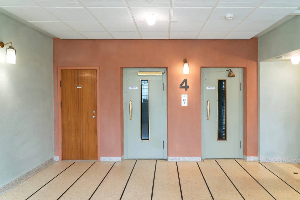 To lysegrønne heisdører og en tredør er plassert i en rosa vegg merket 4 i en godt opplyst, ryddig oppgang med stripete gulv.