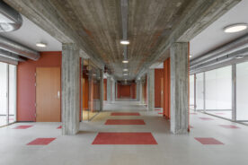 En moderne, tom gang har betongsøyler, røde dører, glassvegger og et mønstret gulv med røde og grå fliser under taklamper.