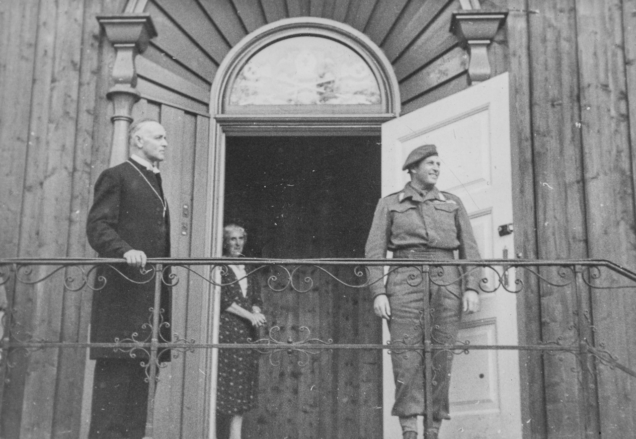 Tre personer står utenfor en trebygning; en mann i dress, en kvinne i kjole i døråpningen og en person i militæruniform på en veranda med åpen dør.