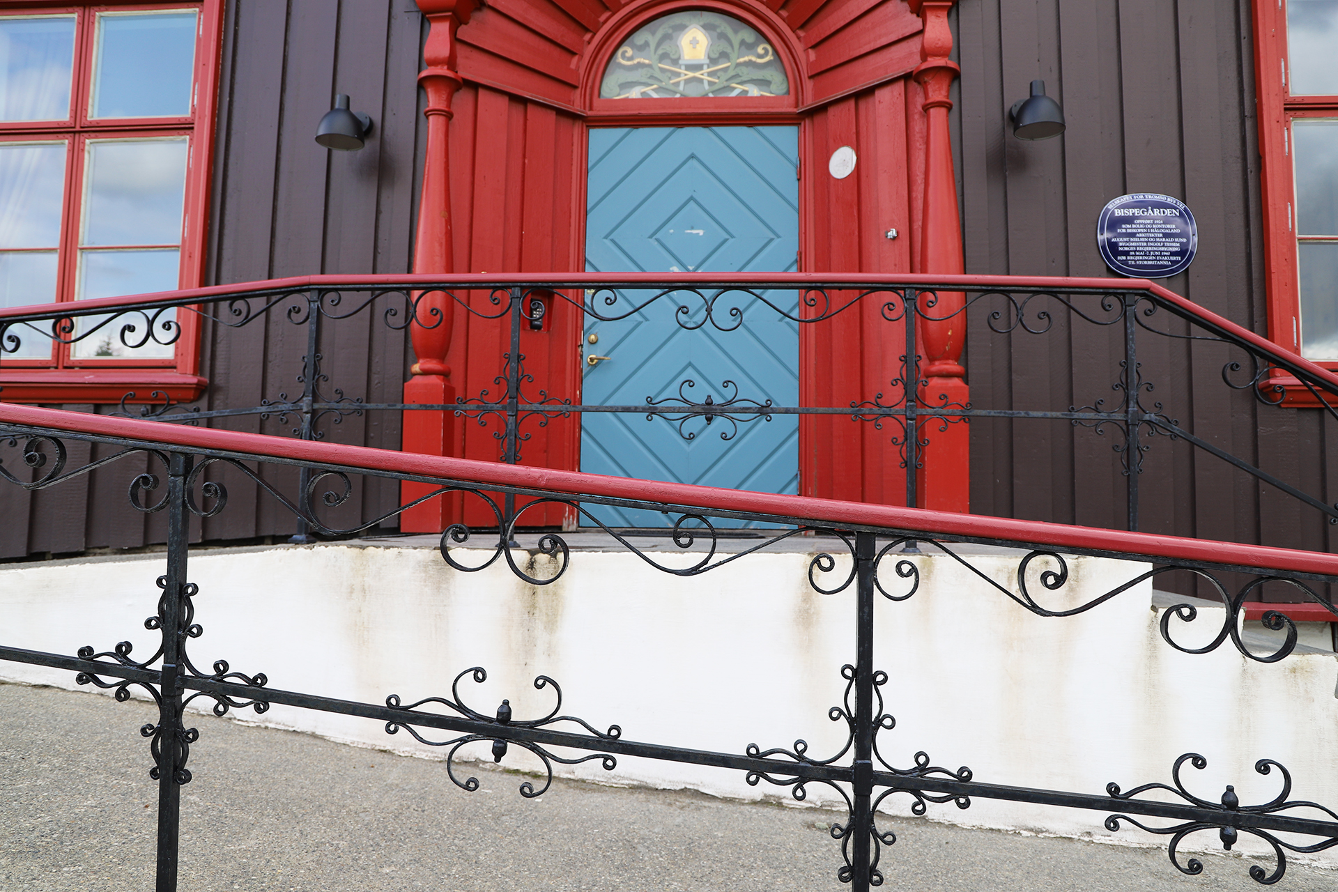 En blå dør med diamantmønster er plassert i en brun og rød trebygning, med et dekorativt, svart metallrekkverk foran.