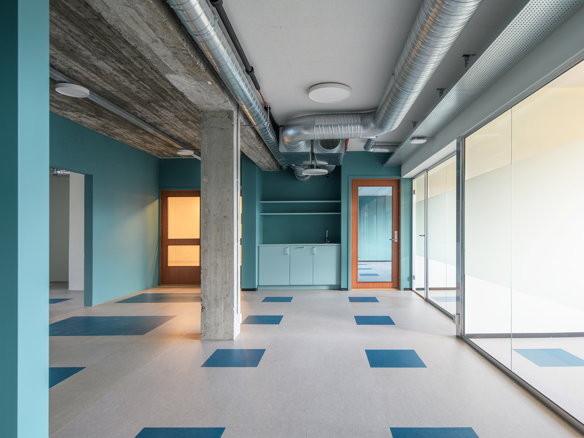 Moderne kontorinteriør med blågrønne vegger, eksponert betong og ventilasjonskanaler, skillevegger i glass, tredører og blåmønstrede fliser på gulvet.