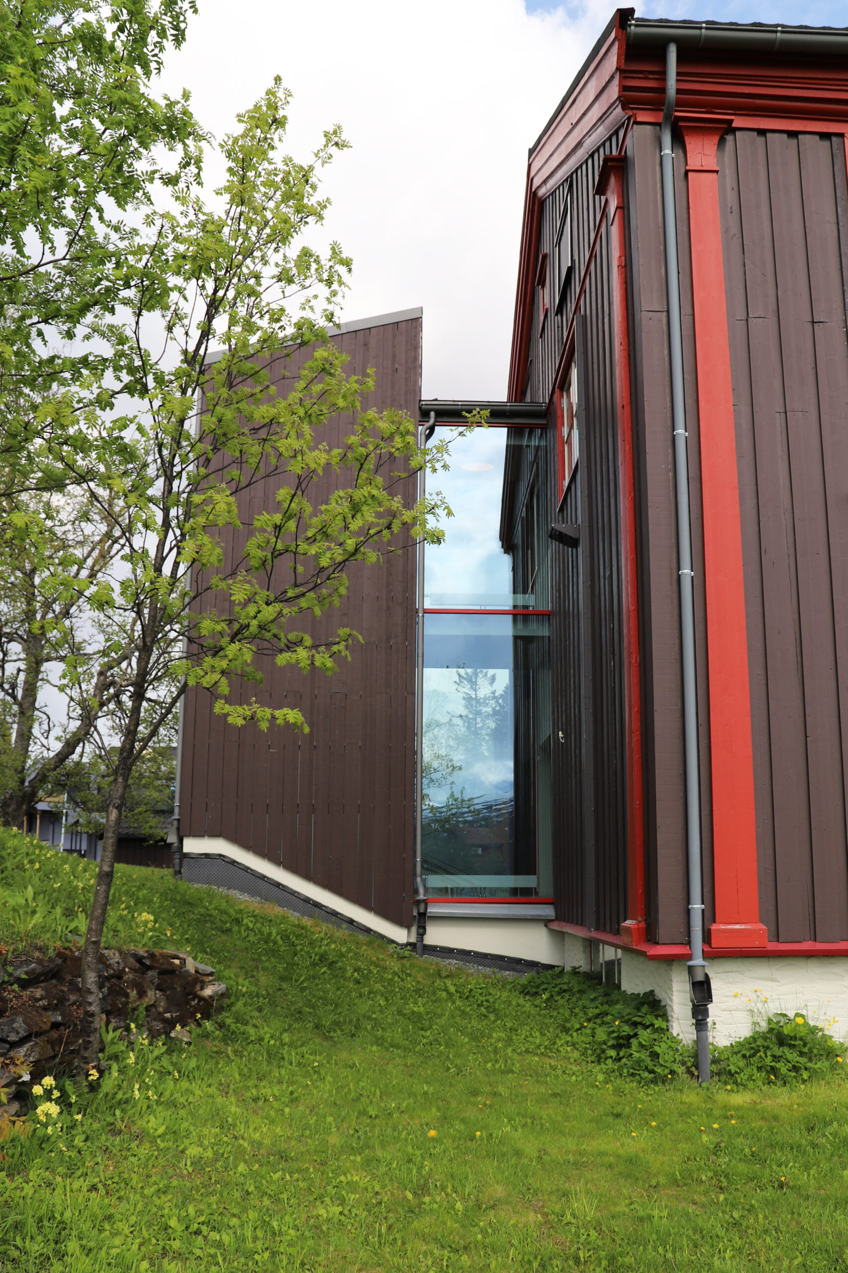En moderne bygning med mørk trekledning og rød listverk har et høyt vindusparti i glass; i forgrunnen ser man et lite tre og grønt gress.