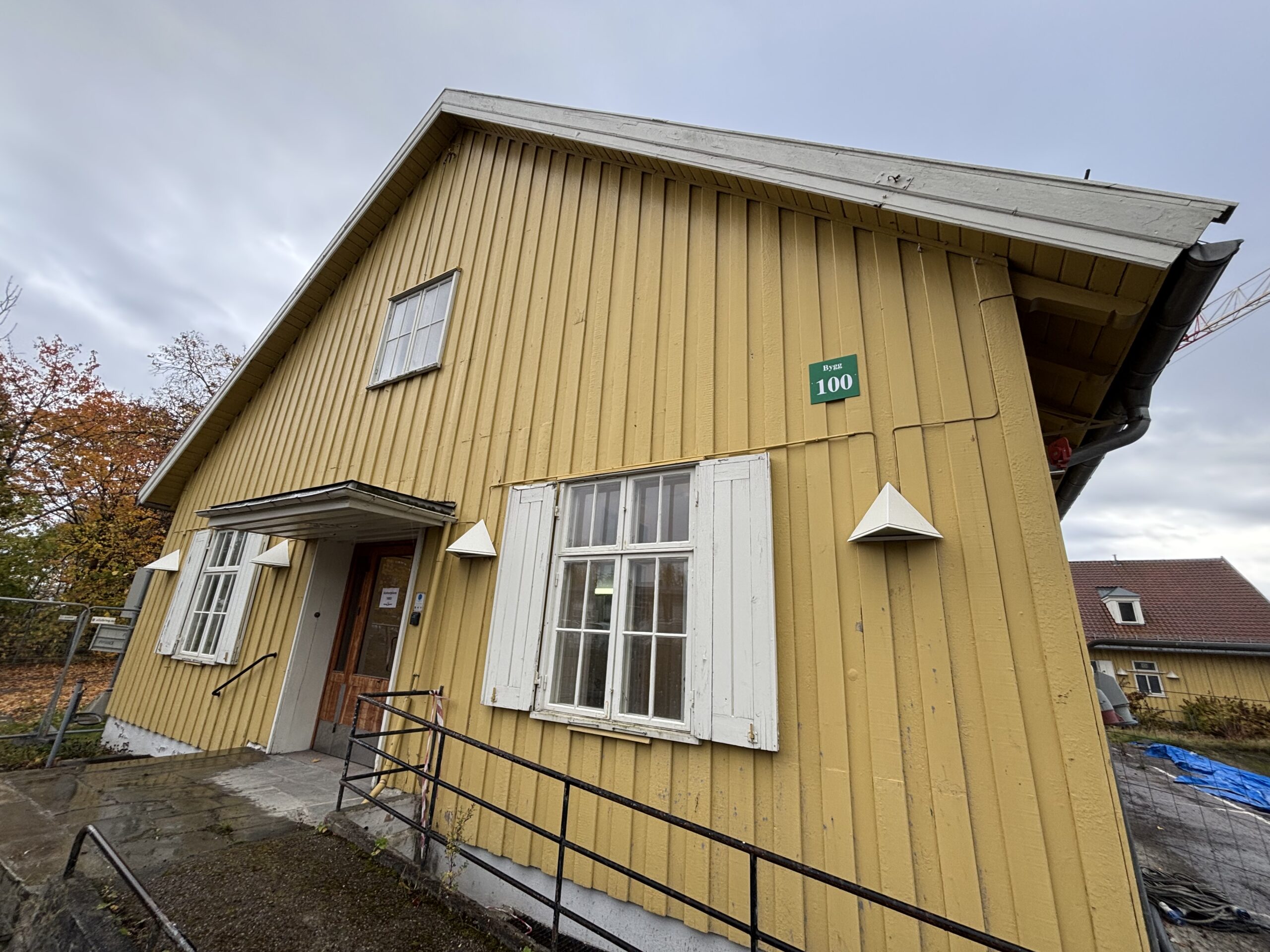 Et gult trehus med hvite kanter, rampeadkomst og et skilt med tallet 100 på veggen; overskyet himmel og høsttrær i bakgrunnen.