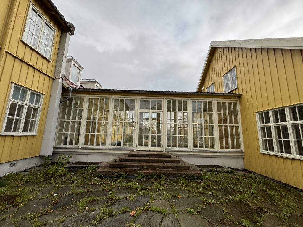 En gårdsplass med steinbelegg og ugress, omgitt av gule trebygninger og en sentral glassveranda med trapper som fører til doble dører.