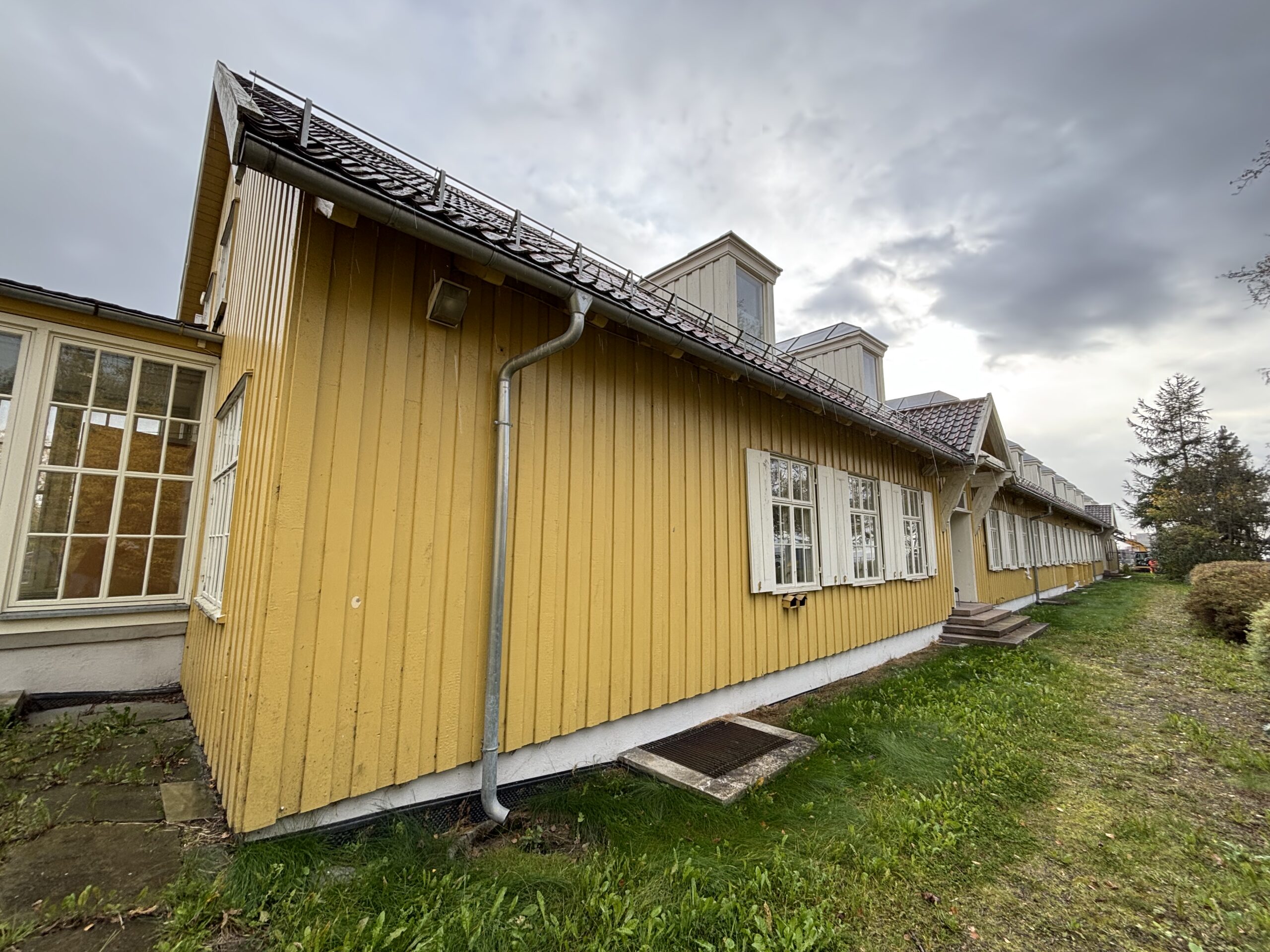 En lang, gul trebygning med hvite vinduer og skrått tak, fotografert fra siden på en overskyet dag. Gress og en sti løper langs bygningen.