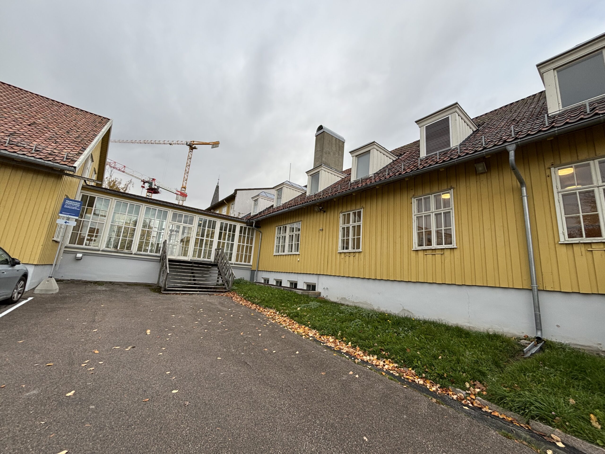 En gul trebygning med hvitkledde vinduer, forbundet med en gangbro av glass, med en kran synlig i bakgrunnen under en overskyet himmel.