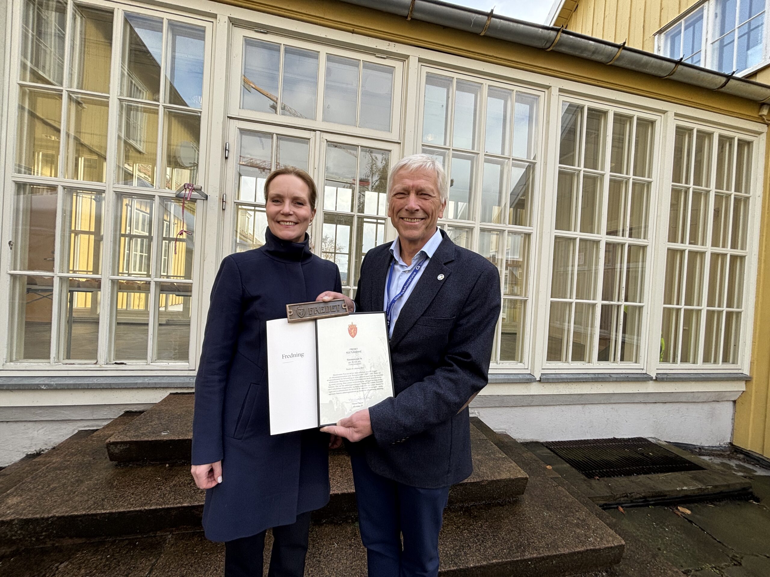 To personer står utenfor en gul bygning, smiler og holder et offisielt dokument oppslått mellom seg.