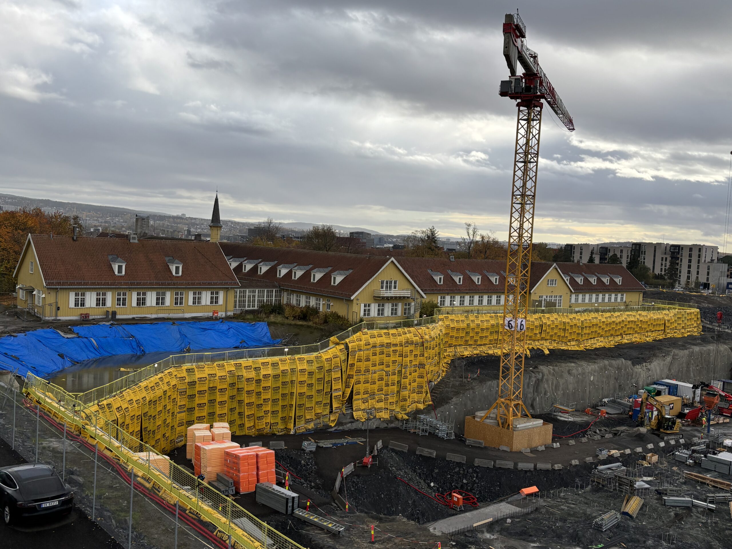 En byggeplass med en stor gul kran, gule sikkerhetsbarrierer, stabler med materialer og bolighus i bakgrunnen under en overskyet himmel.