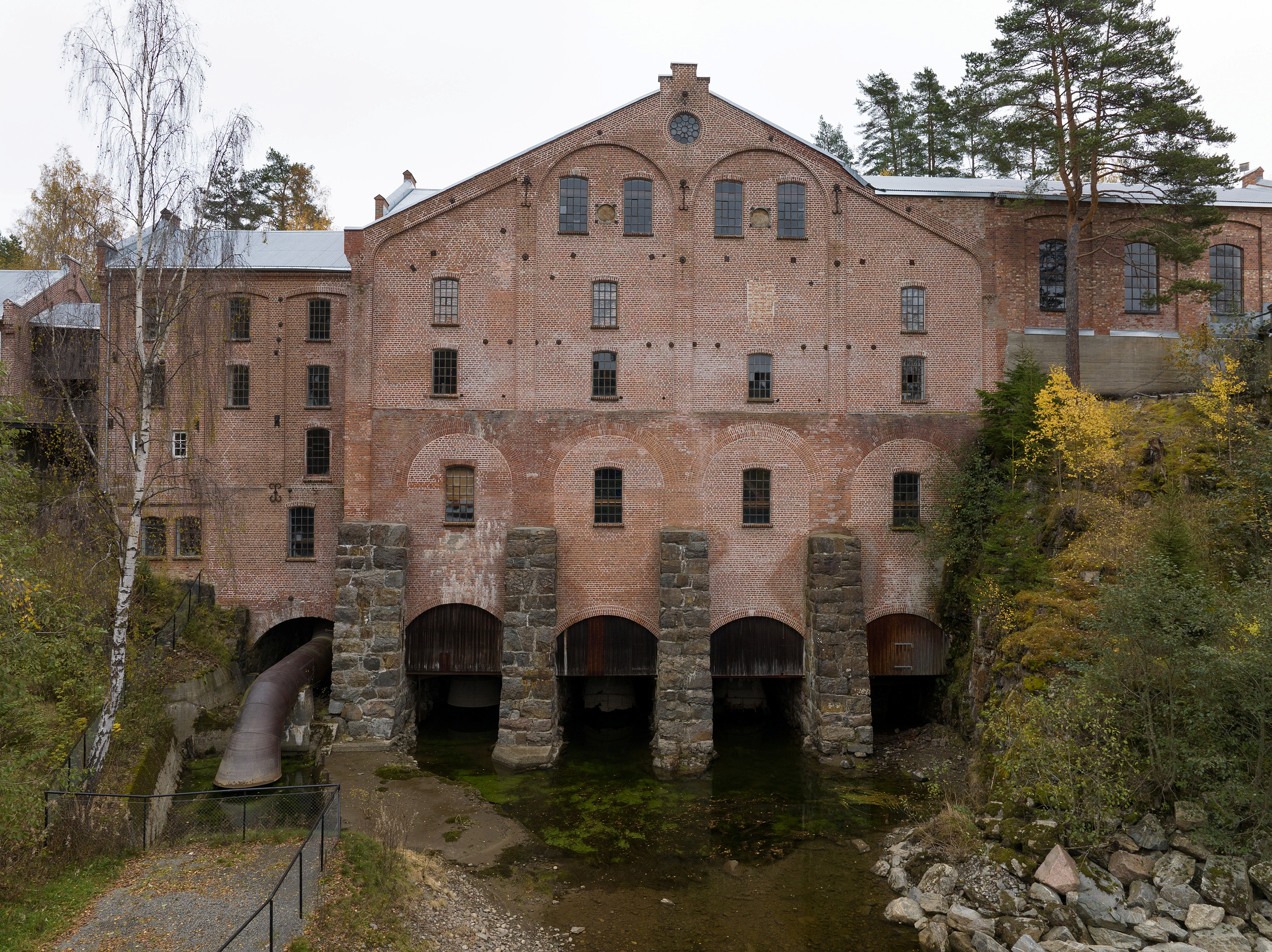En stor, gammel industribygning i teglstein med flere vinduer står over steinbuer over en grunn elv, omgitt av høsttrær.