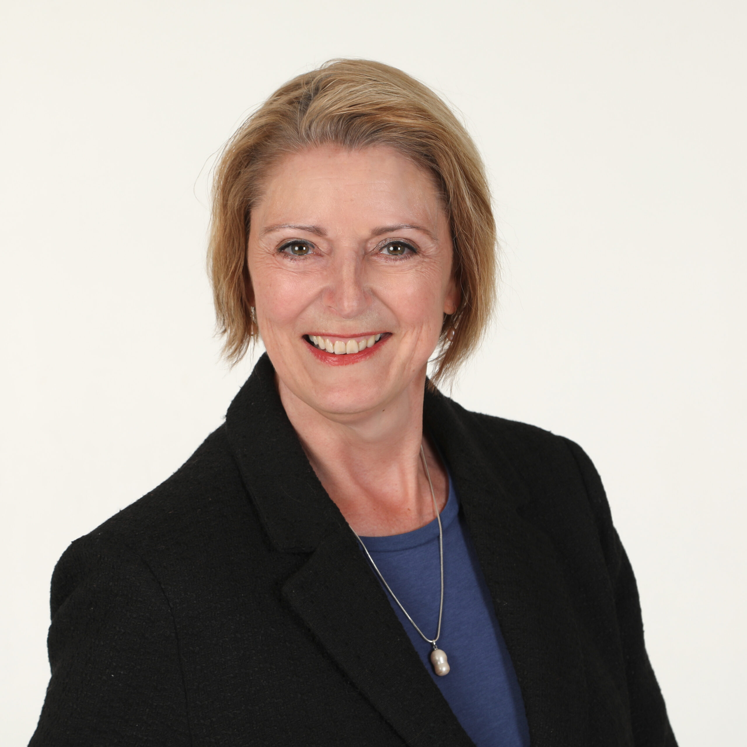 En kvinne med kort, blondt hår iført svart blazer, blå topp og halskjede med anheng smiler til kameraet mot en hvit bakgrunn.