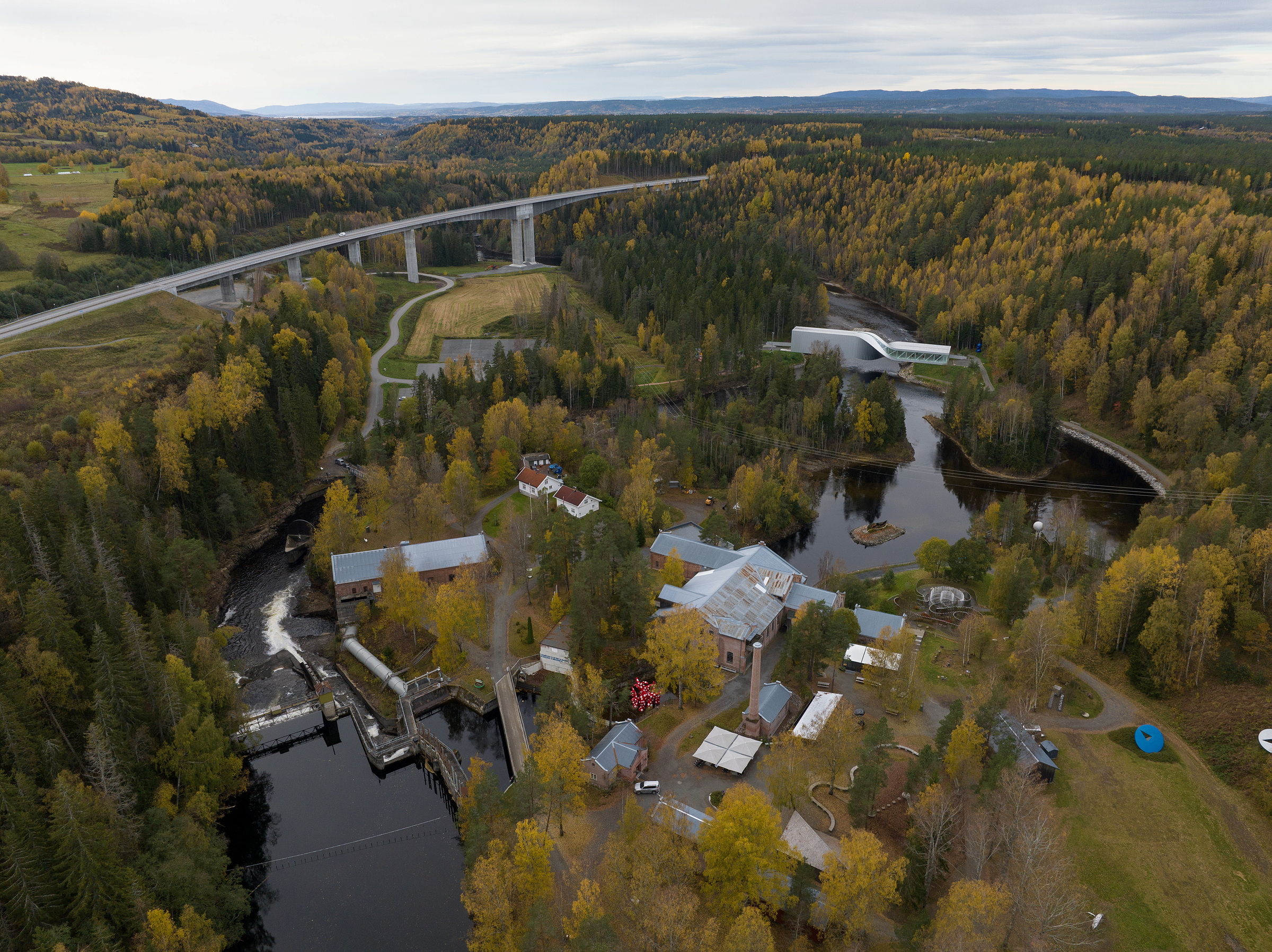 Flyfoto av Kistefosmuseet med elv, bygninger, en demning, høstløvverk og en stor bro som krysser landskapet.