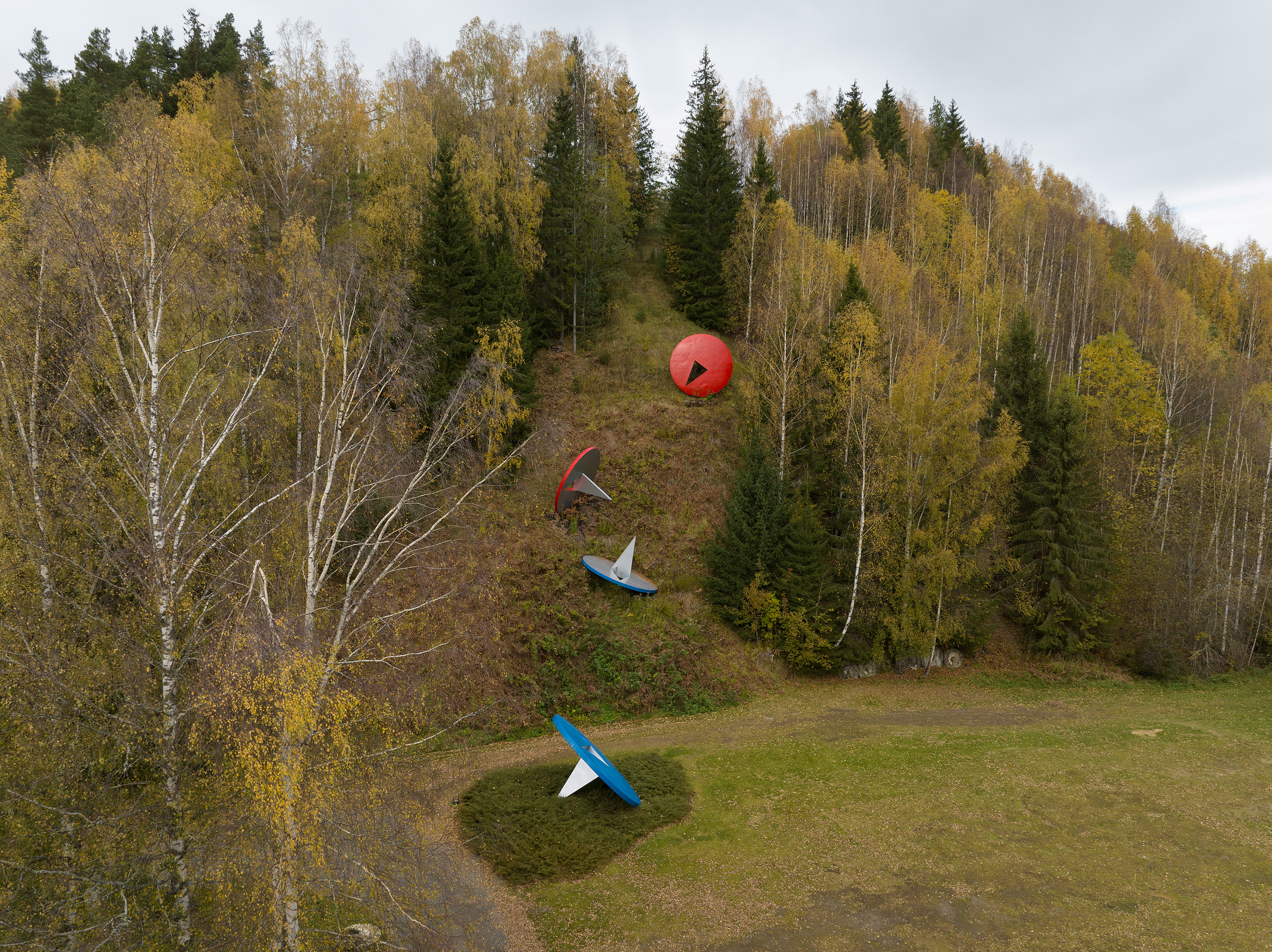 Fire fargerike objekter som ligner parabolantenner og en stor rød kule er plassert på en gressbakke omkranset av høsttrær under en overskyet himmel.