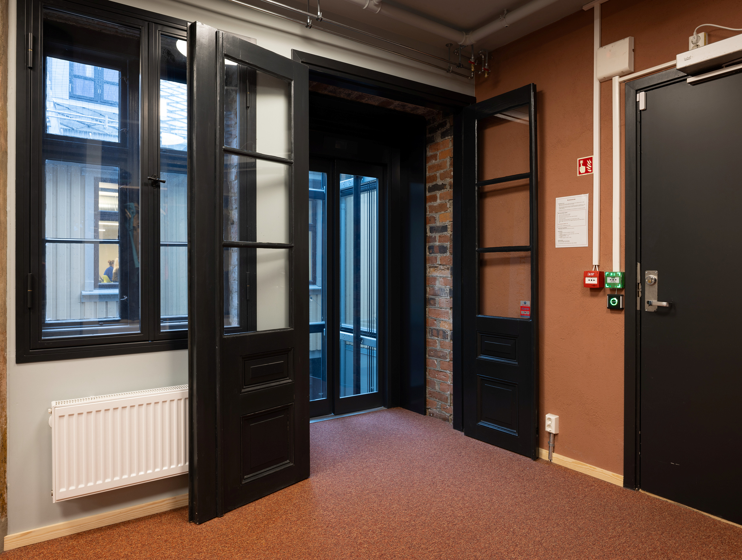 En gang med åpne, svarte glassdører, brune vegger, nødknapper, en radiator og utsikt mot en bygning utenfor.