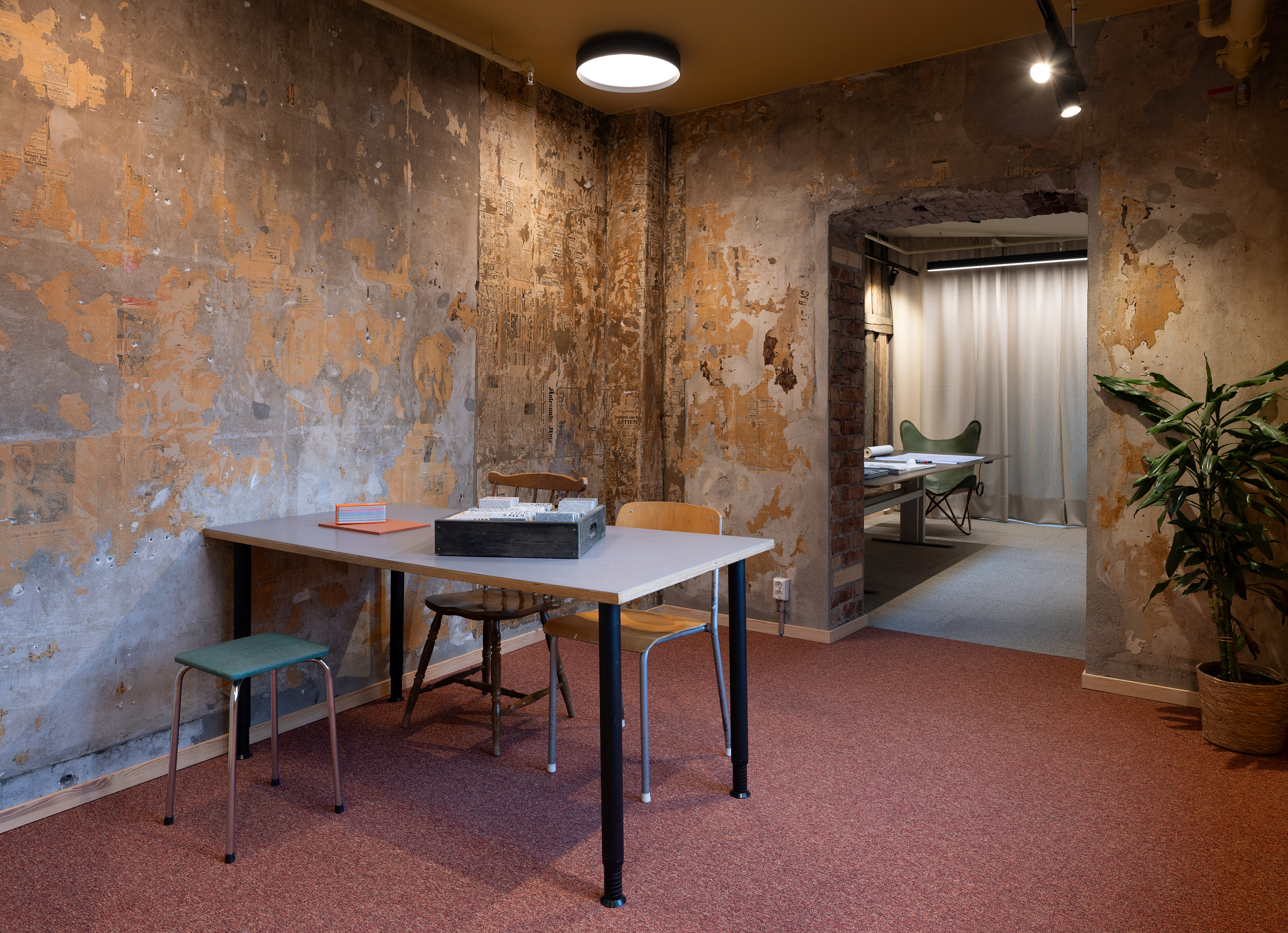 Et minimalistisk rom med avskallede vegger, et hvitt bord omgitt av stoler som ikke passer sammen, og en potteplante; et tilstøtende rom med en seng er synlig gjennom en åpen døråpning.