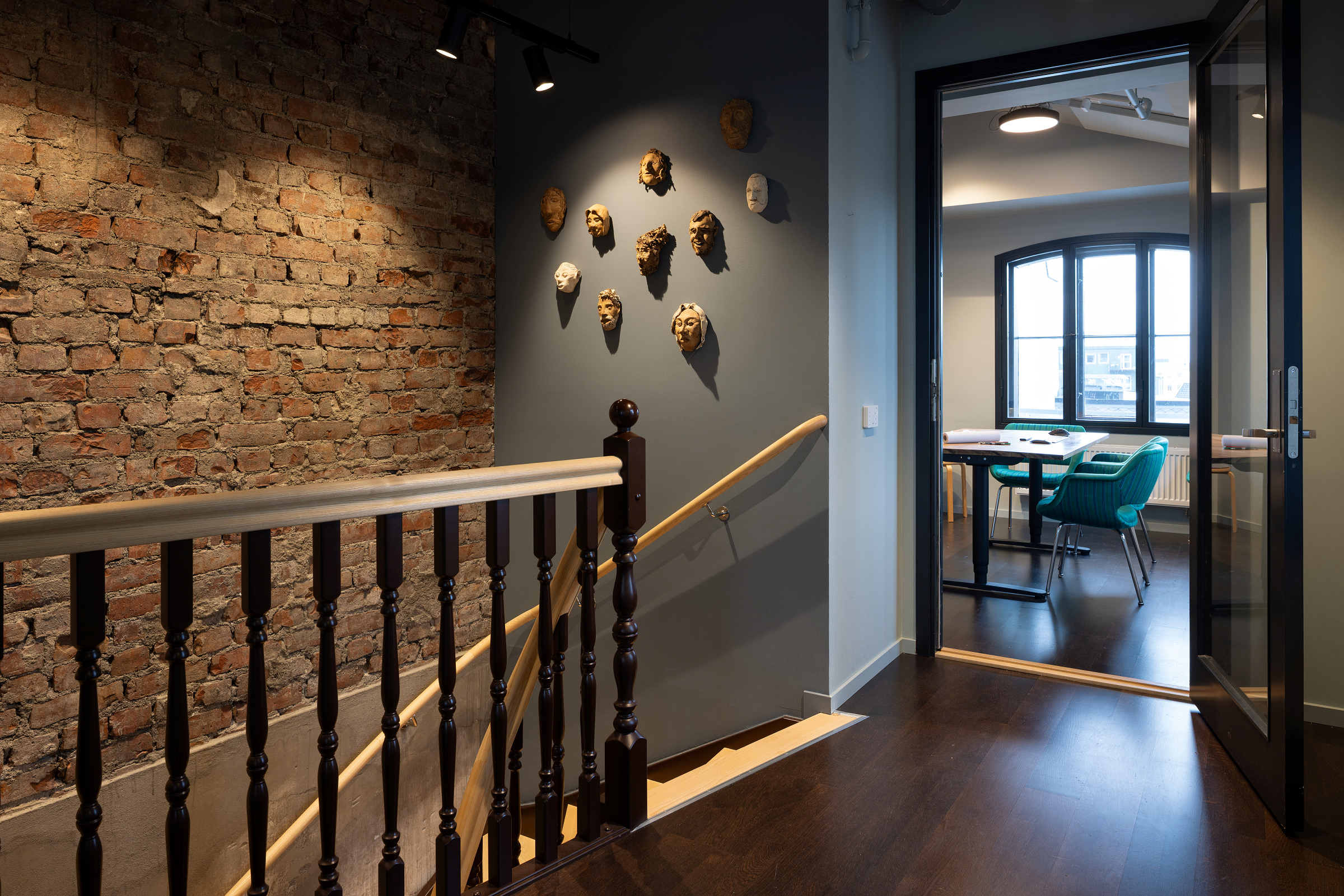 En trapp med rekkverk i tre, eksponert murvegg og dekorative masker på veggen fører til et kontor med bord, blågrønne stoler og et stort vindu.