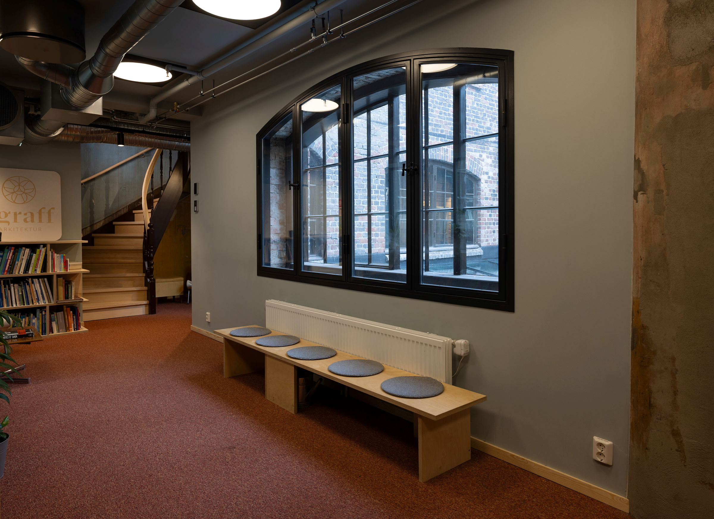 Et moderne interiør med en trebenk og runde, grå puter under et stort vindu, rødt teppegulv, bokhyller og en vindeltrapp i bakgrunnen.
