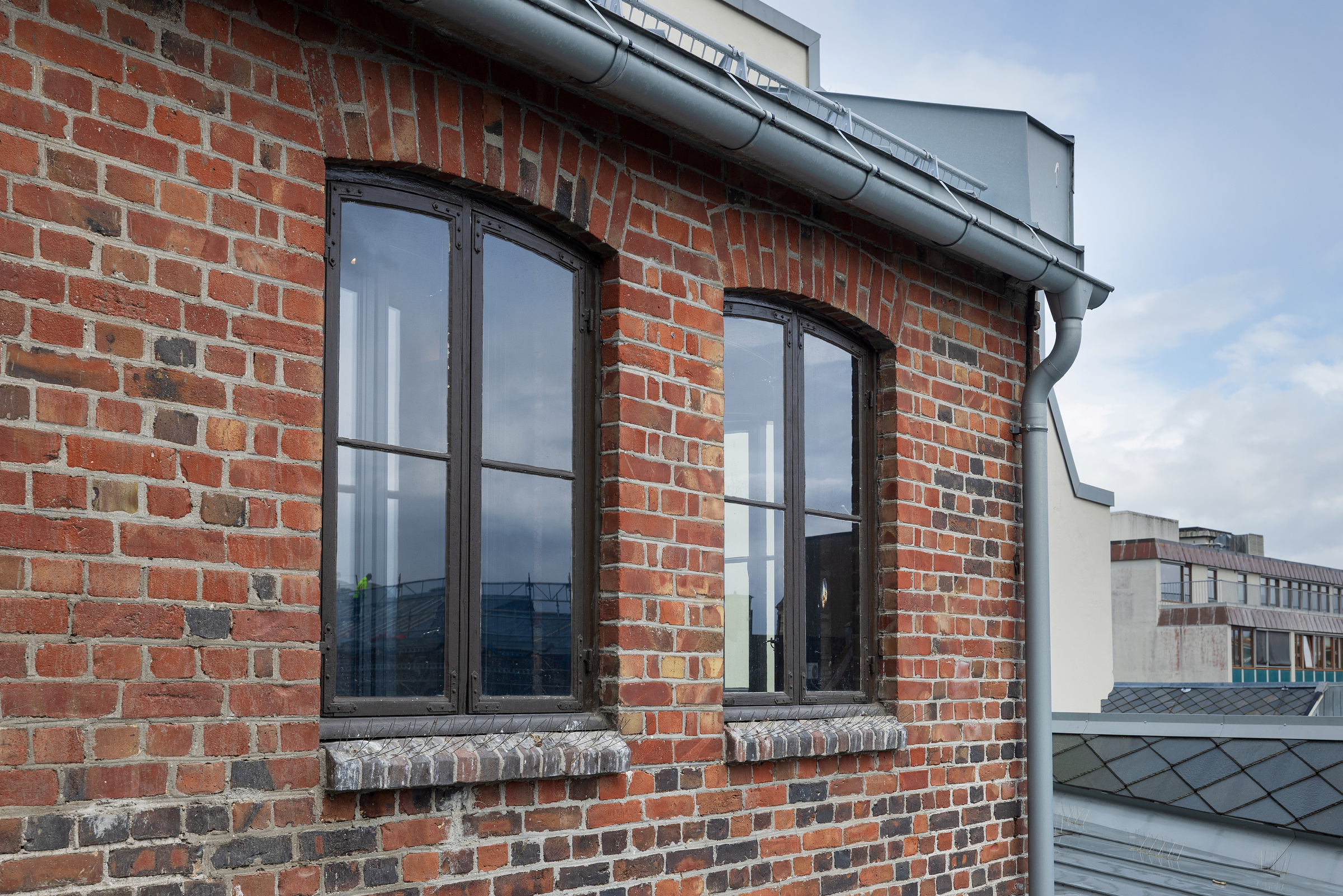 To buede vinduer i en rød murvegg, med en grå takrenne av metall over og andre bygninger synlige i bakgrunnen.