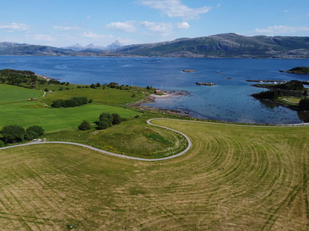 En svingete vei svinger seg gjennom grønne åkrer ved siden av et stille vann, med fjellene synlige i bakgrunnen under en delvis overskyet himmel.