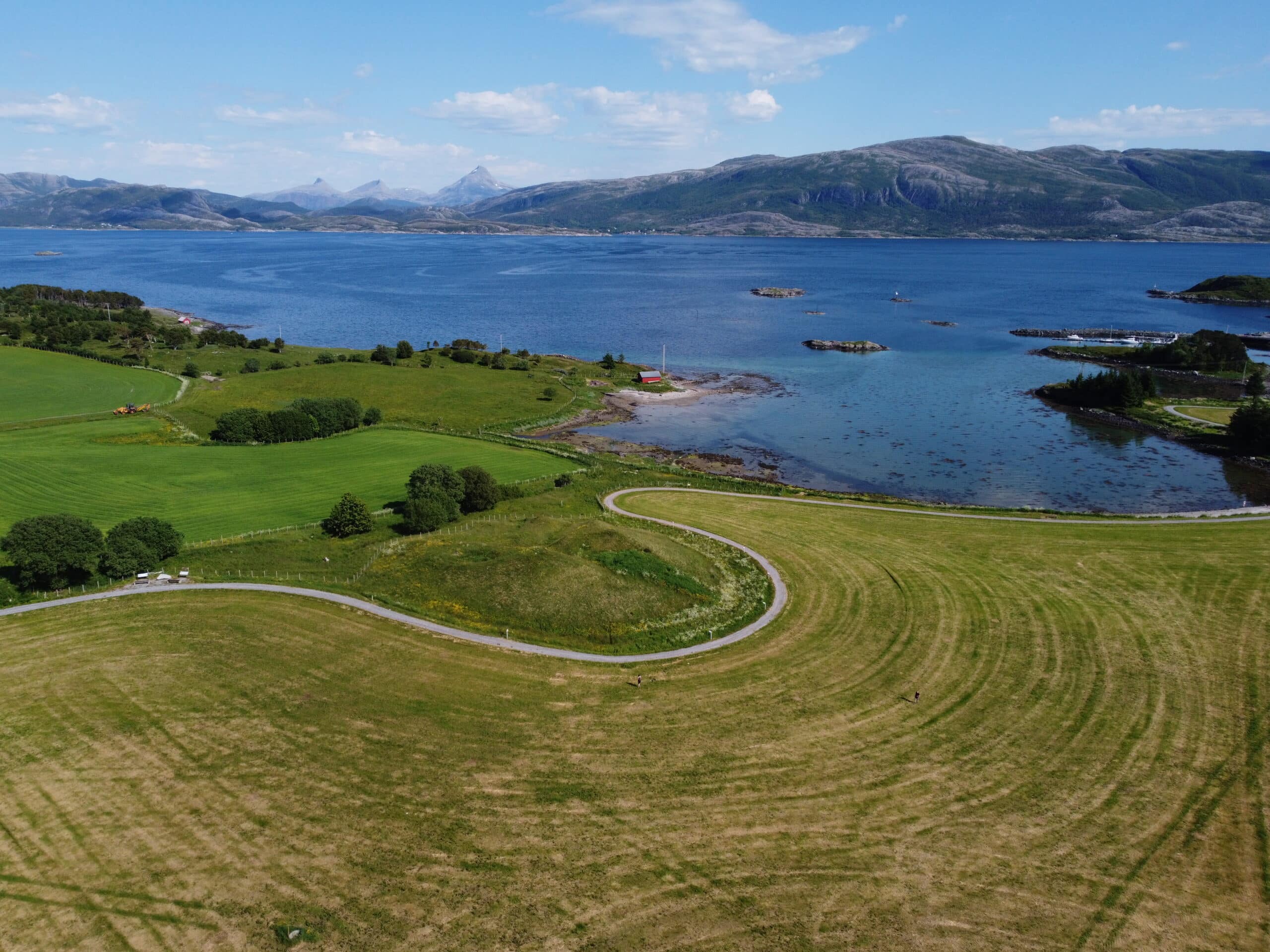 En svingete vei svinger seg gjennom grønne åkrer ved siden av et stille vann, med fjellene synlige i bakgrunnen under en delvis overskyet himmel.