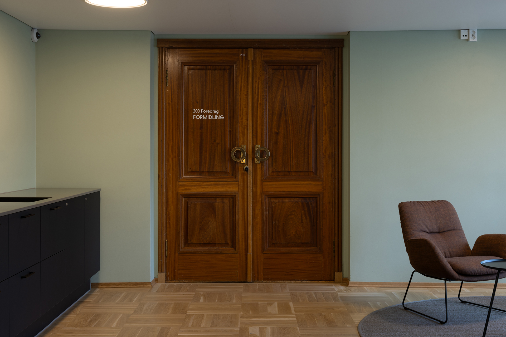 Et par lukkede dobbeltdører i tre merket 207 Föreläsn FORMIDLING i et minimalistisk rom med en brun stol og svarte skap.