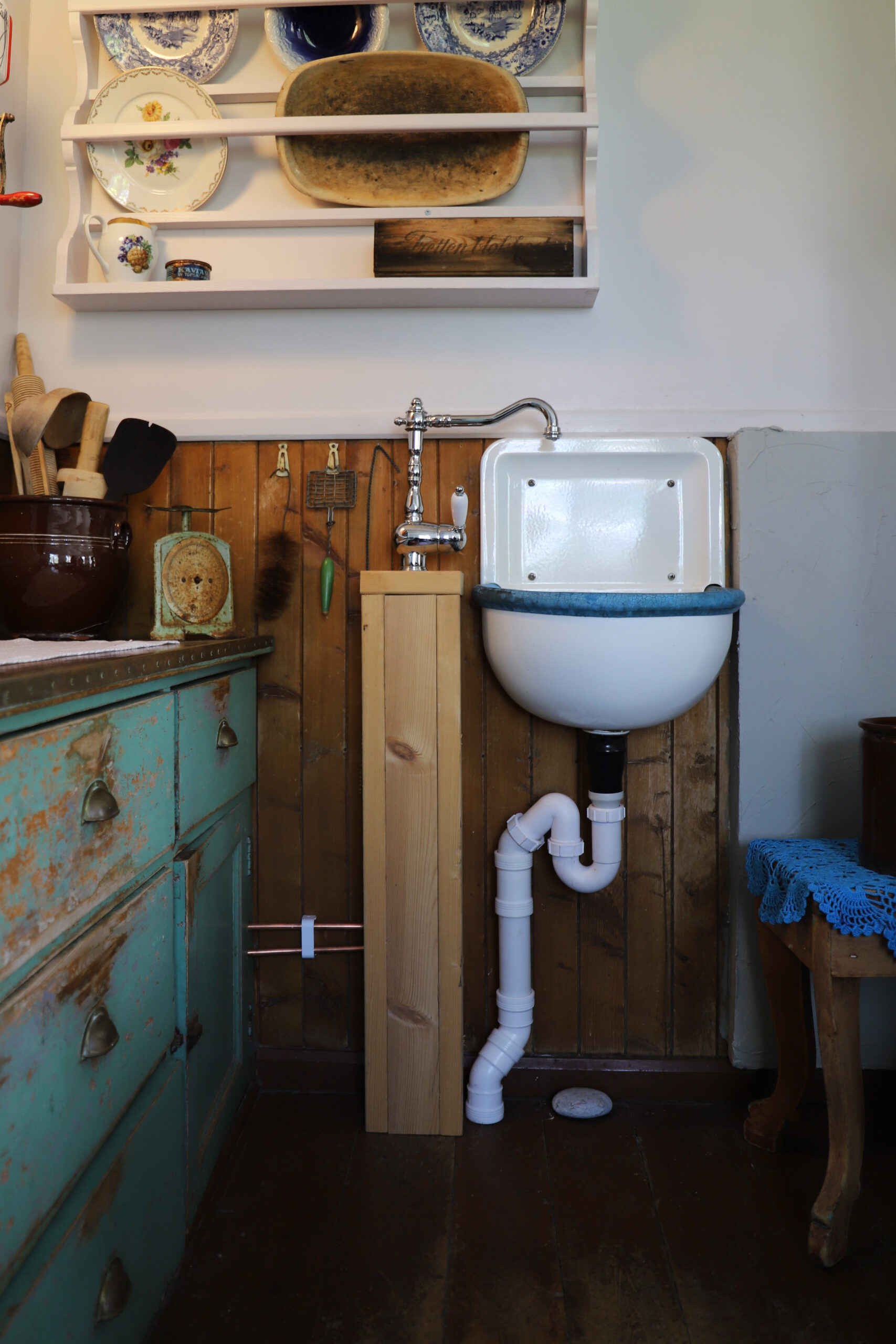 Et lite vintagekjøkken med veggmontert vask, synlige rør, bakplater i tre, hyller med servise og et slitt grønt skap.