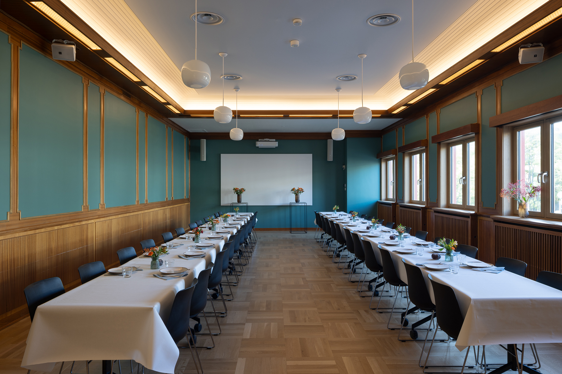 Et konferanserom med to langbord dekket til et måltid, svarte stoler, tregulv, blågrønne vegger, store vinduer og en whiteboardtavle foran.