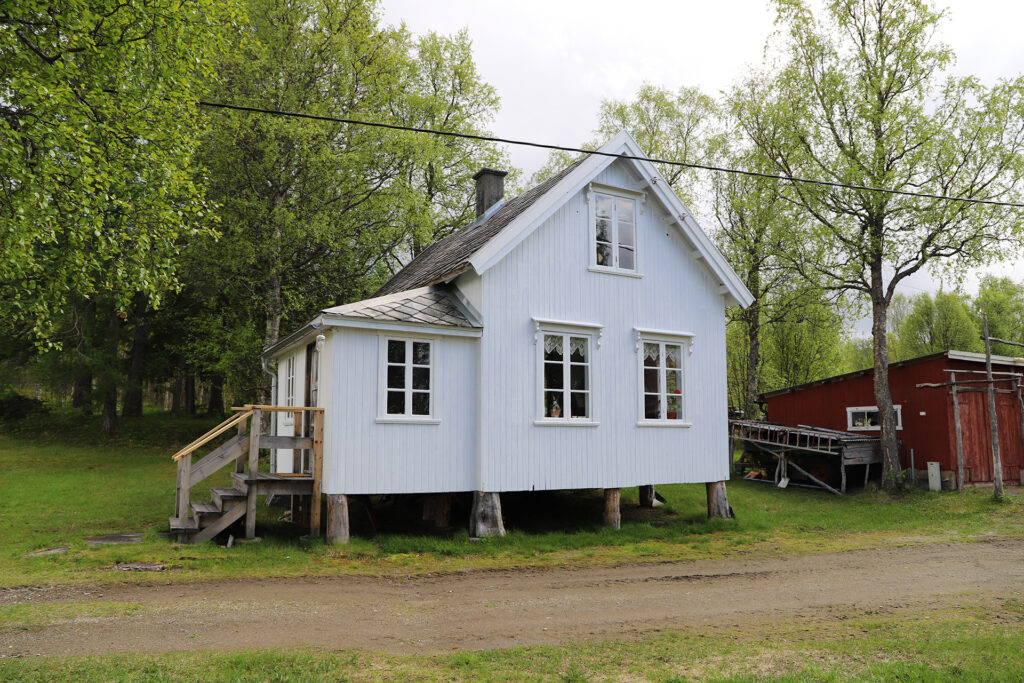 Et lite, hvitt trehus på stylter med veranda, omgitt av grønne trær og gress, står ved siden av et rødt uthus.