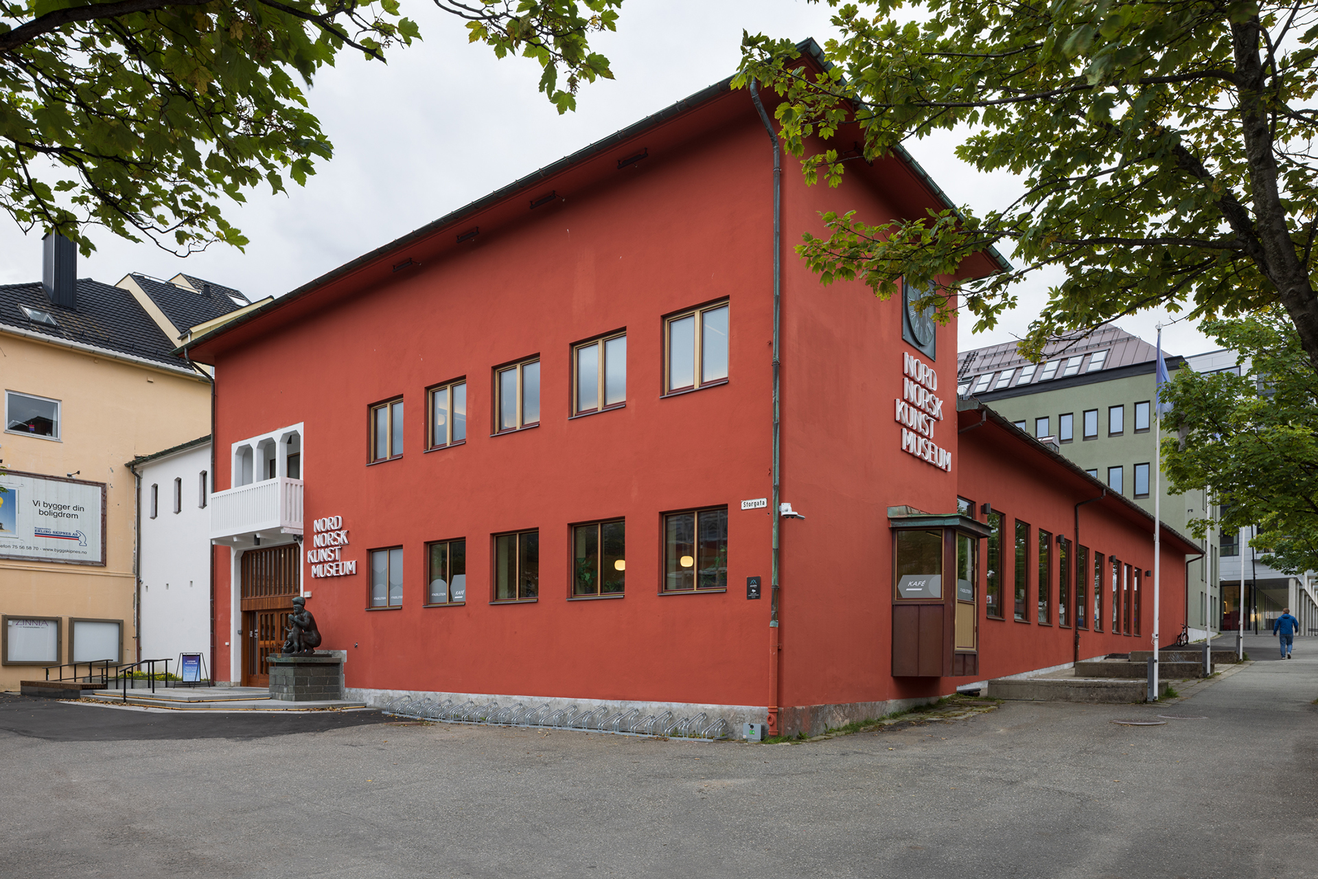 En to-etasjers rød bygning med flere vinduer, merket Nord Norsk Kunstmuseum, ligger på et gatehjørne med trær som delvis rammer inn scenen.