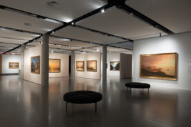 Et moderne kunstgalleri med malerier på hvite vegger, opplyst av taklamper, og svarte, runde benker på et polert betonggulv.