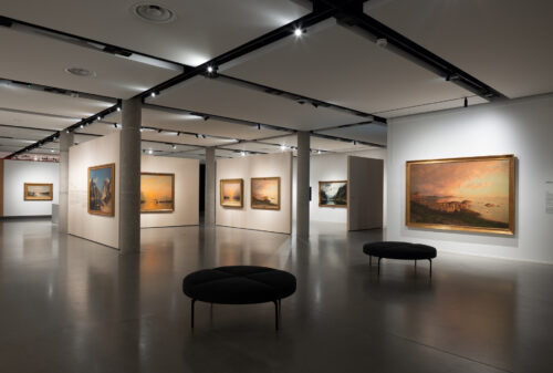 Et moderne kunstgalleri med malerier på hvite vegger, opplyst av taklamper, og svarte, runde benker på et polert betonggulv.