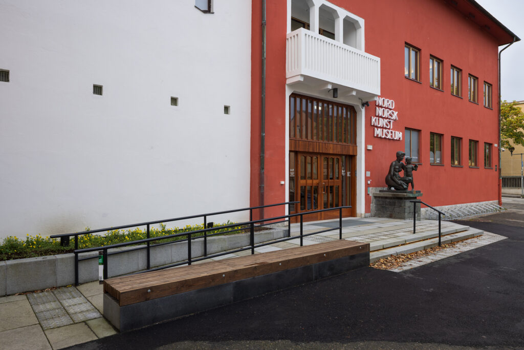 Inngangspartiet til Nordnorsk Kunstmuseum med en statue ved døren, røde og hvite yttervegger og en trebenk i forgrunnen.