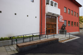 Inngangspartiet til Nordnorsk Kunstmuseum med en statue ved døren, røde og hvite yttervegger og en trebenk i forgrunnen.