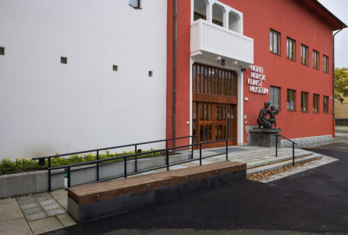 Inngangspartiet til Nordnorsk Kunstmuseum med en statue ved døren, røde og hvite yttervegger og en trebenk i forgrunnen.