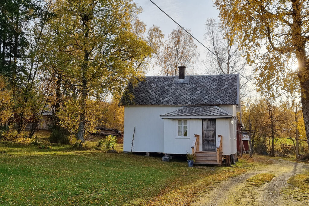 Et lite, hvitt trehus med takspon og en liten veranda, omgitt av høsttrær og en gresskledd hage under en klar himmel.